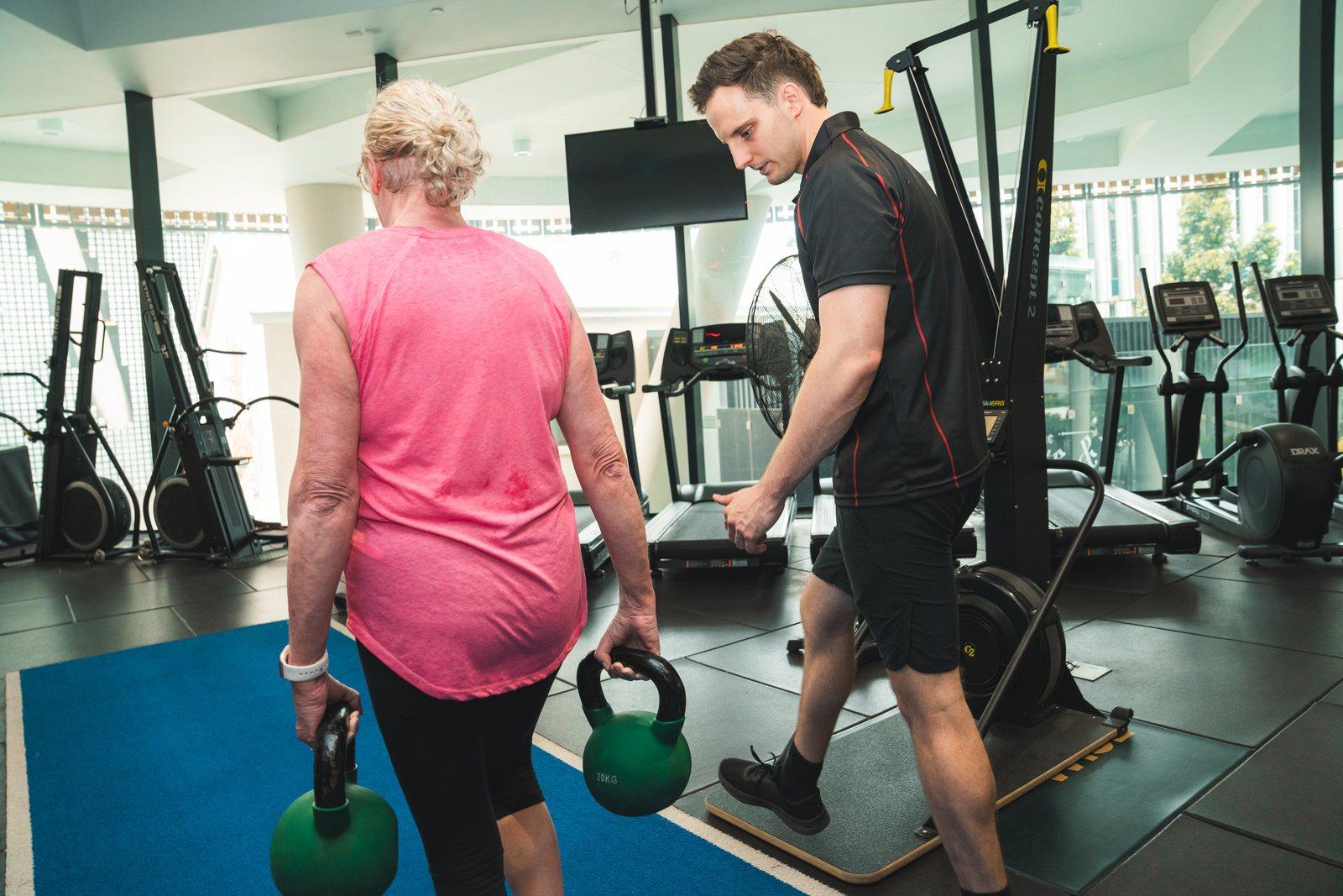 A Catalyst client in her sixties carrying two 20kg kettlebells across the gym floor while Nick walks beside her